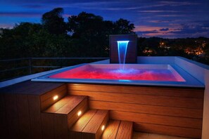 Ein Whirlpool mit Wasserfall, umgeben von Holzstufen und einem Geländer, vor einer Dämmerungshimmelslinie.