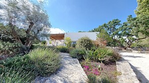 Un bâtiment moderne, auquel on accède par un chemin bordé de pierres, et qui est entouré de différentes plantes et d’arbres.
