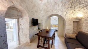 A stone-walled room with a dining table, chairs, a sofa, and a TV mounted on the wall.