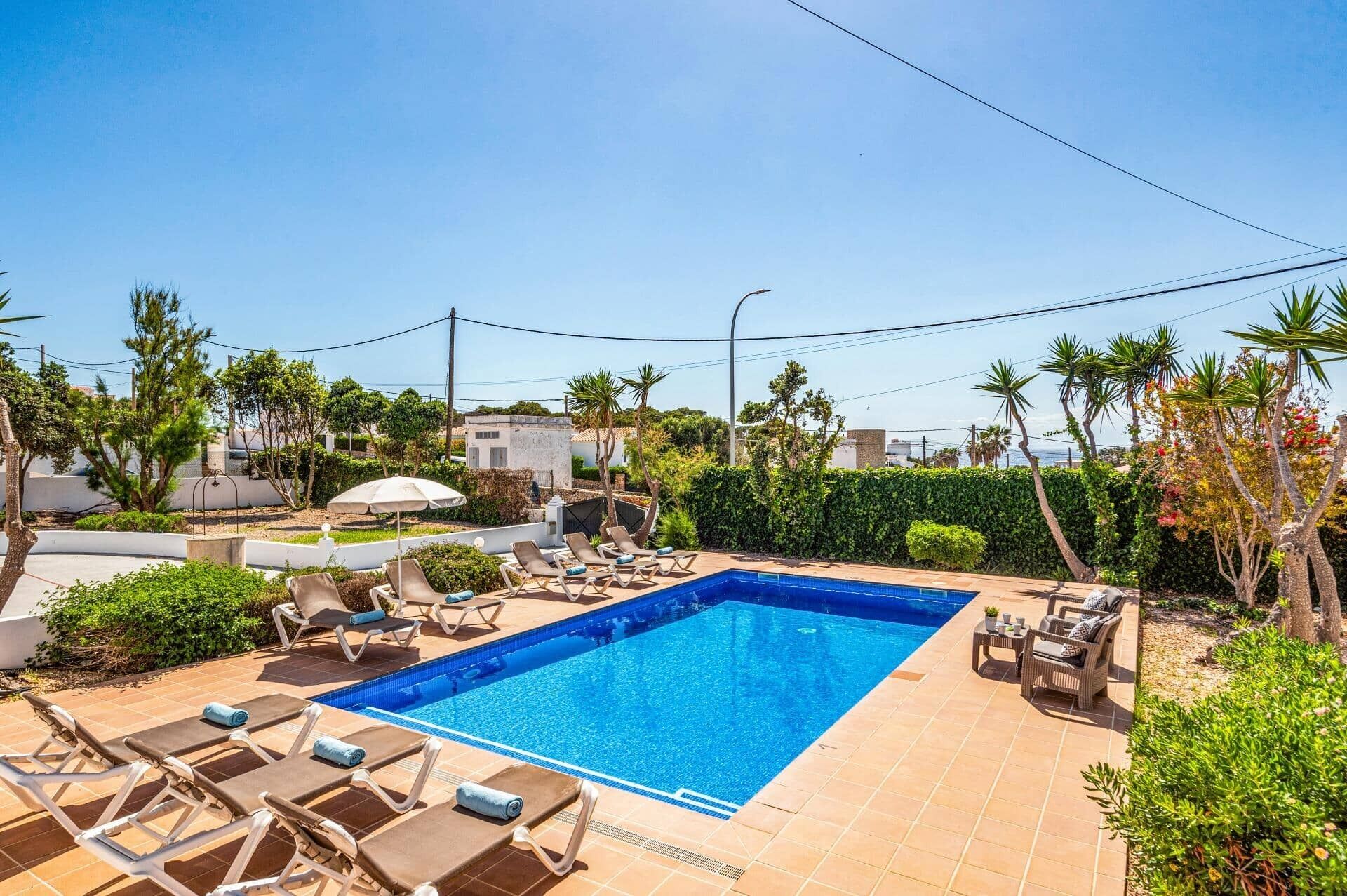 A swimming pool with lounge chairs and a small table, surrounded by greenery and a clear sky.