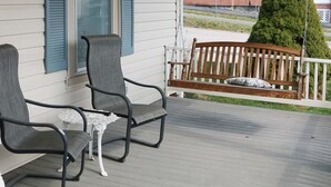 A covered porch with two chairs and a small table, a wooden bench, and a view of a brick building in the background. - Perfect for Everyone - 8-bedrooms - The Future Generations University Guesthouse (Franklin)