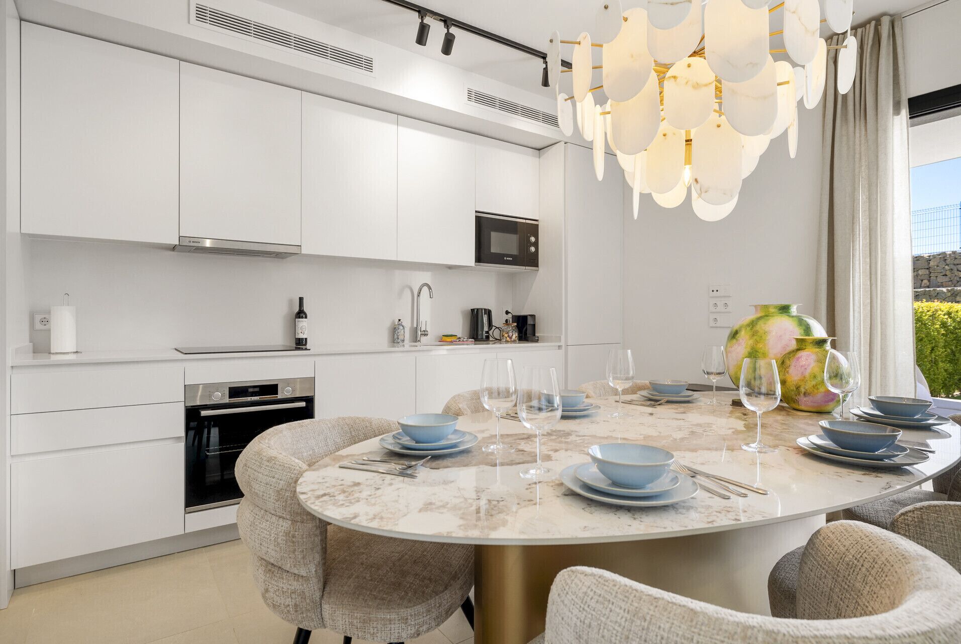 A modern kitchen with white cabinetry, a built-in oven, a microwave, and a dining area with a marble table set for four.