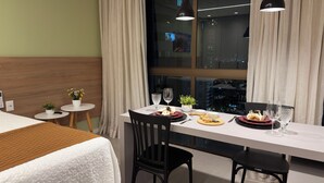 Habitación de hotel con una mesa de comedor preparada para dos personas, una cama con colcha blanca, una pequeña planta en maceta y vistas a la ciudad de noche.