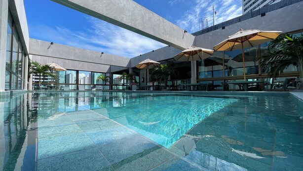 Una piscina interior moderna con agua cristalina, rodeada de sillones de descanso y sombrillas, y grandes ventanales con vistas a la ciudad.