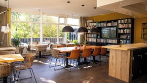 A modern dining area with a central island, surrounded by wicker chairs and tables, with a large bookshelf and a view of greenery outside. - Hotel de Horper Wielen (Kaard)