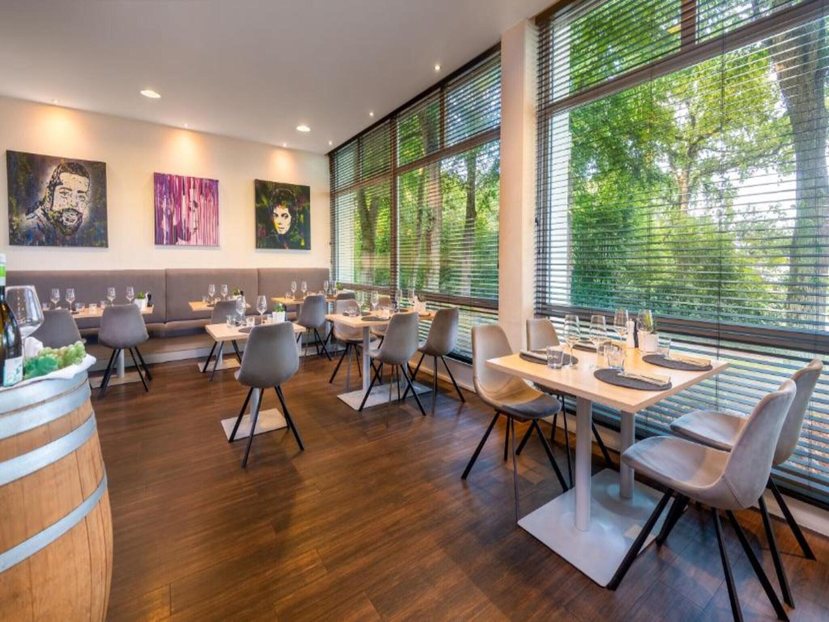A restaurant with a modern interior, featuring wooden flooring, grey upholstered seating, and large windows with blinds.
