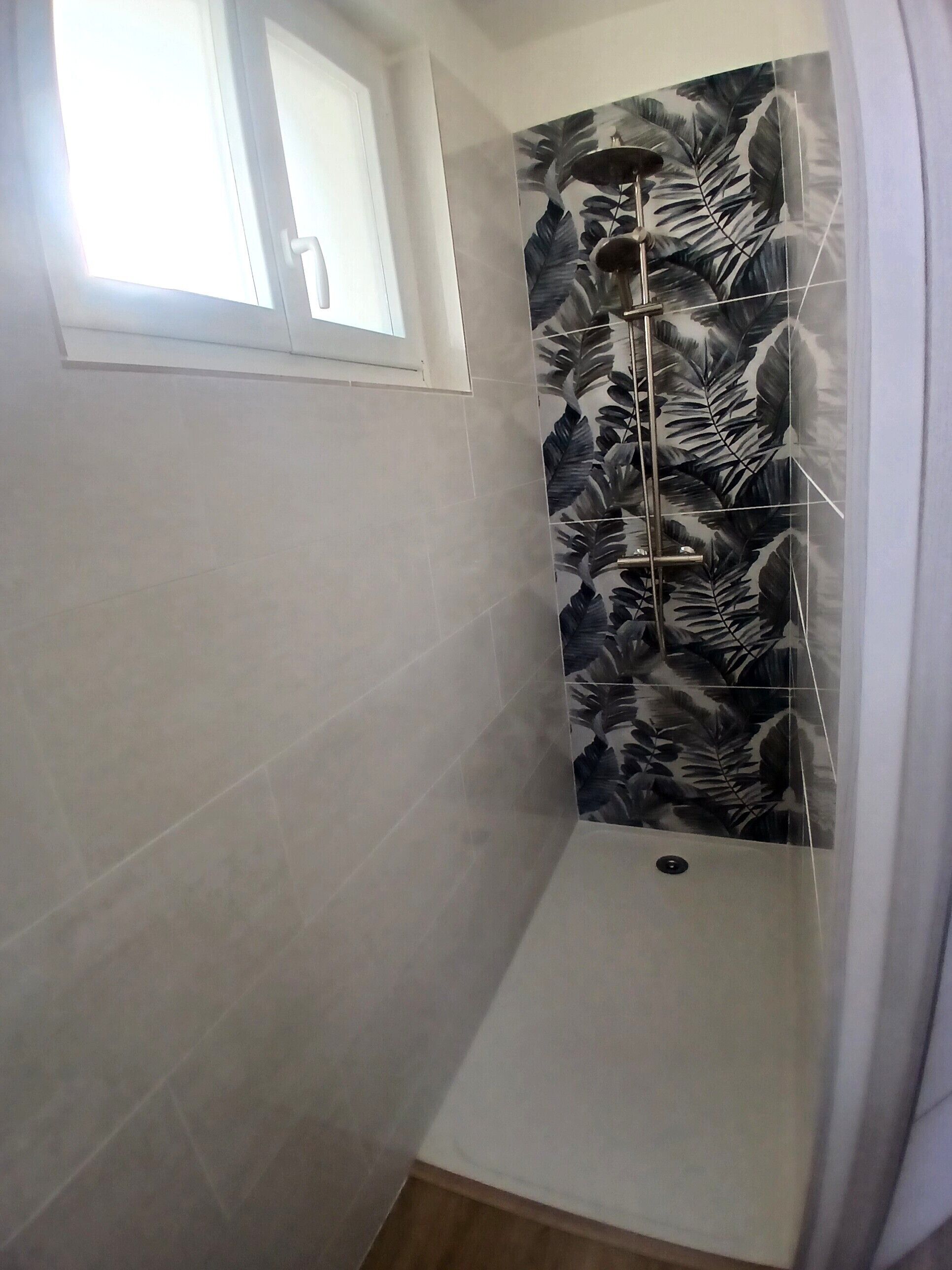 Un bagno con una finestra, una doccia con rivestimento a motivo di foglie e pavimento in legno.