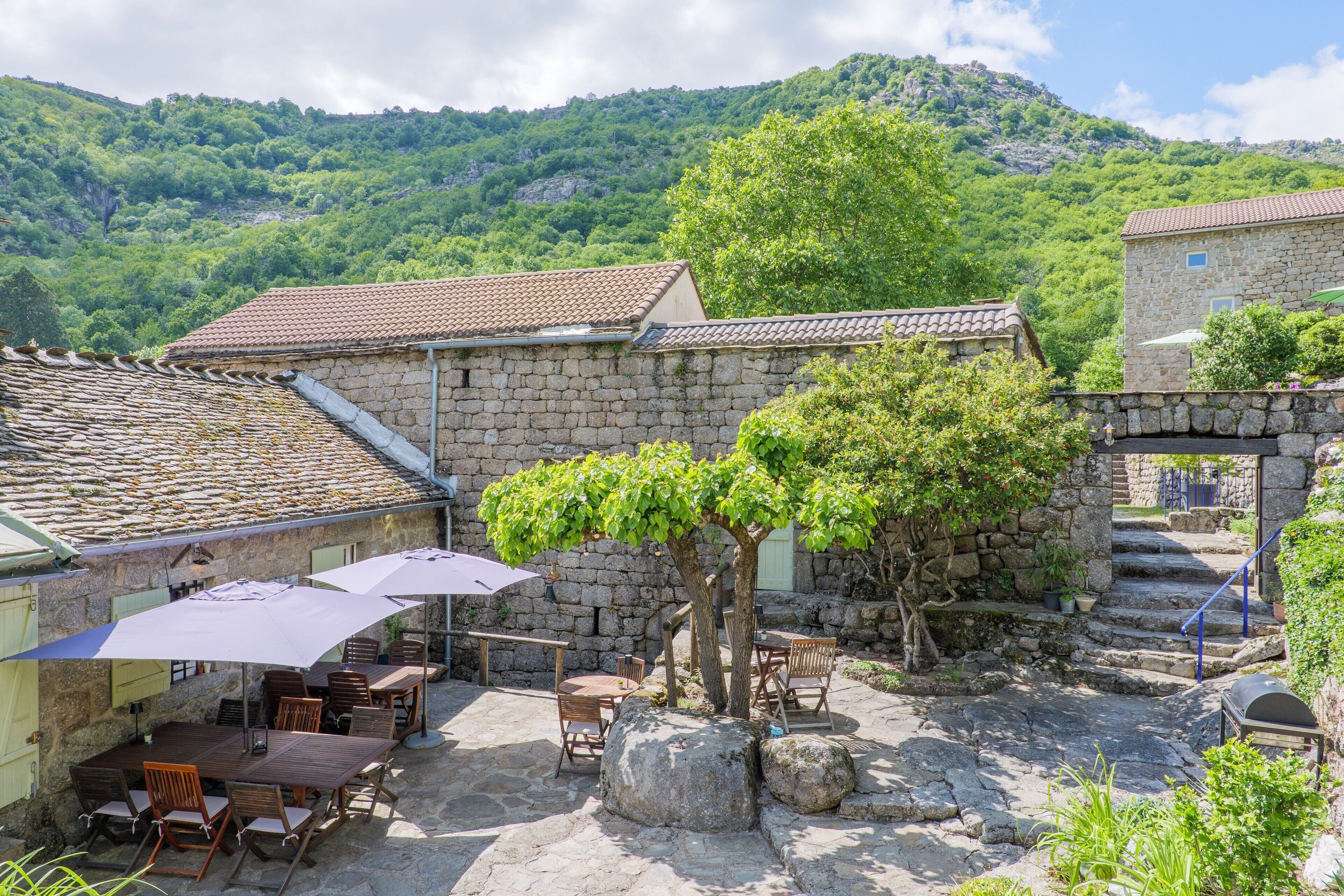 A stone building with a tiled roof, outdoor seating with umbrellas, and a staircase leading to a higher level.