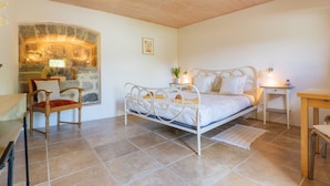 Un dormitorio con cama, una silla, una mesita, una mesita de noche y una pared de piedra.