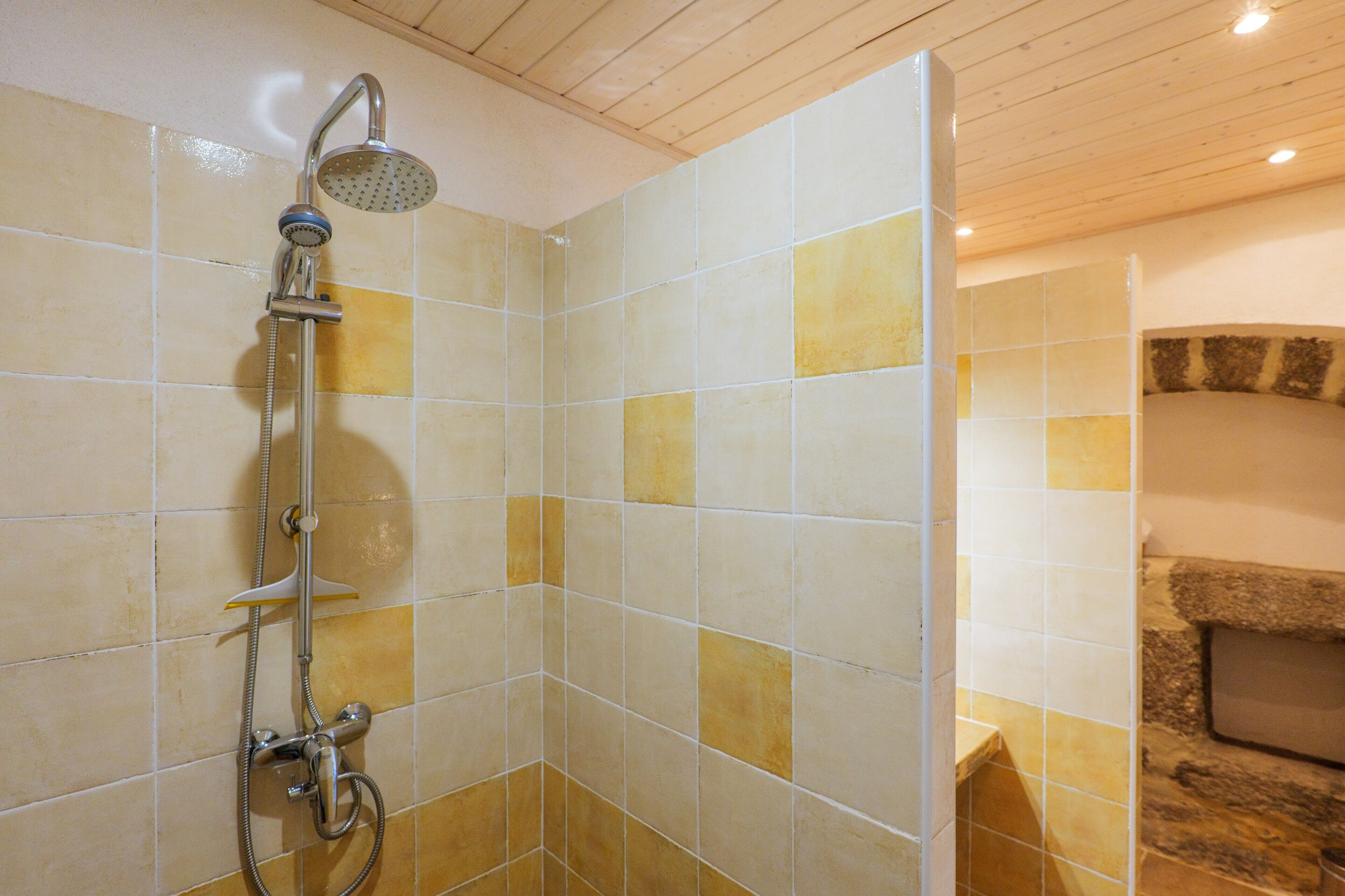 Ducha con cabezal de ducha de mano y grifo empotrado en la pared, paredes con azulejos de diseño variado, combinando baldosas cuadradas y rectangulares, y techo de madera.