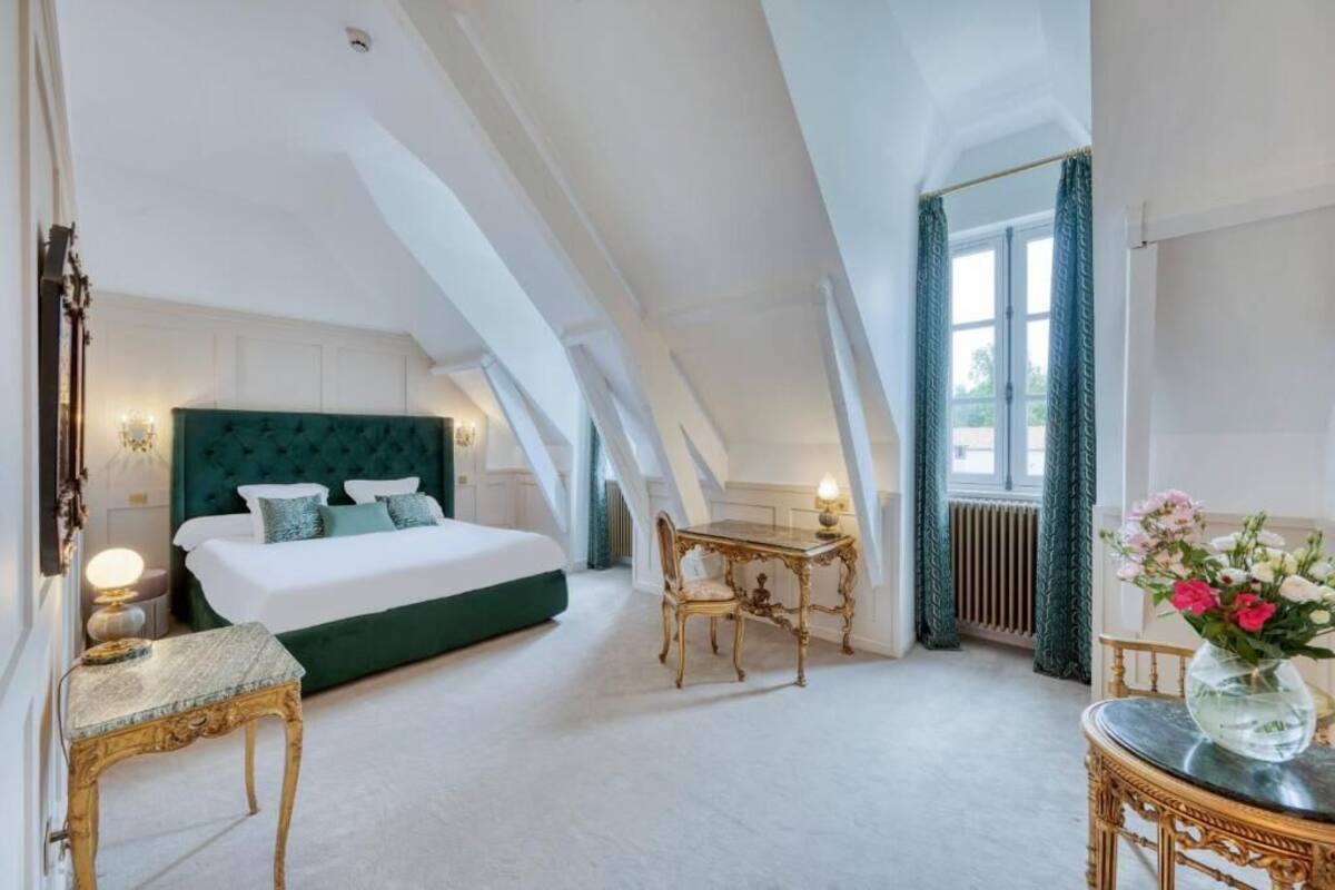 Een slaapkamer met een groot bed, een klein tafeltje, een stoel en een vaas met bloemen.
