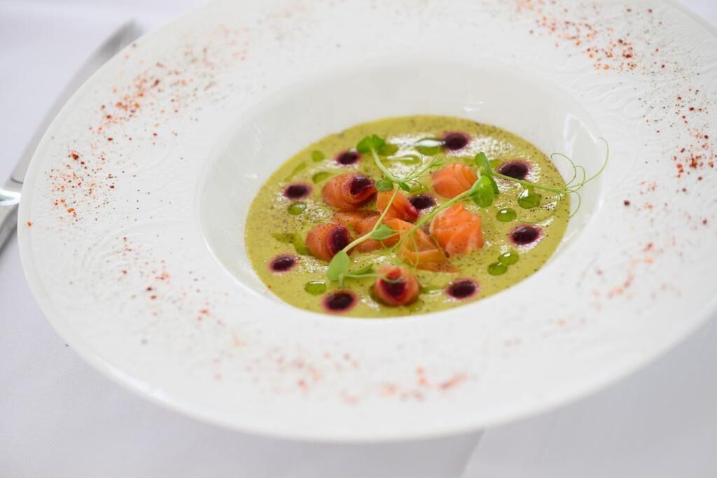 A white plate with a green soup, garnished with herbs and salmon pieces.