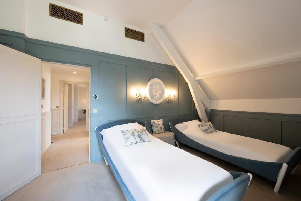 A bedroom with two single beds, a wall-mounted light fixture, and a door leading to another room.