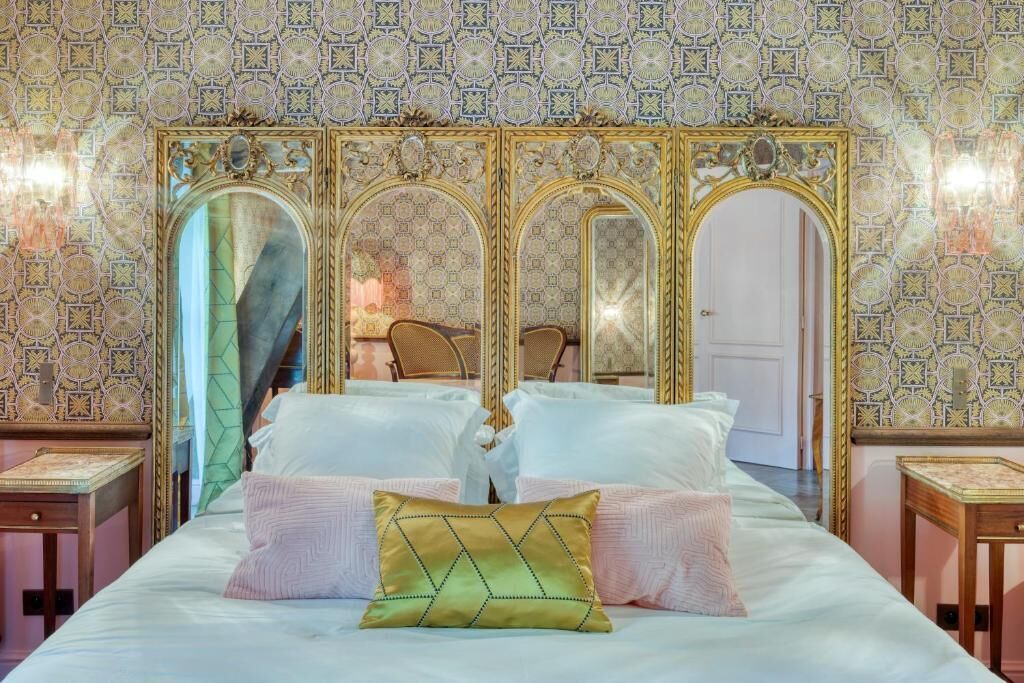 Una cama con múltiples almohadas, un espejo de marcos ornamentados y un papel pintado con diseño.