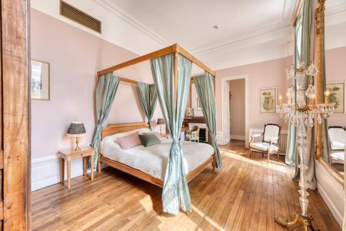 Een kamer met een hemelbed met gordijnen, een kroonluchter en een spiegel, met houten vloeren en een roze wand.