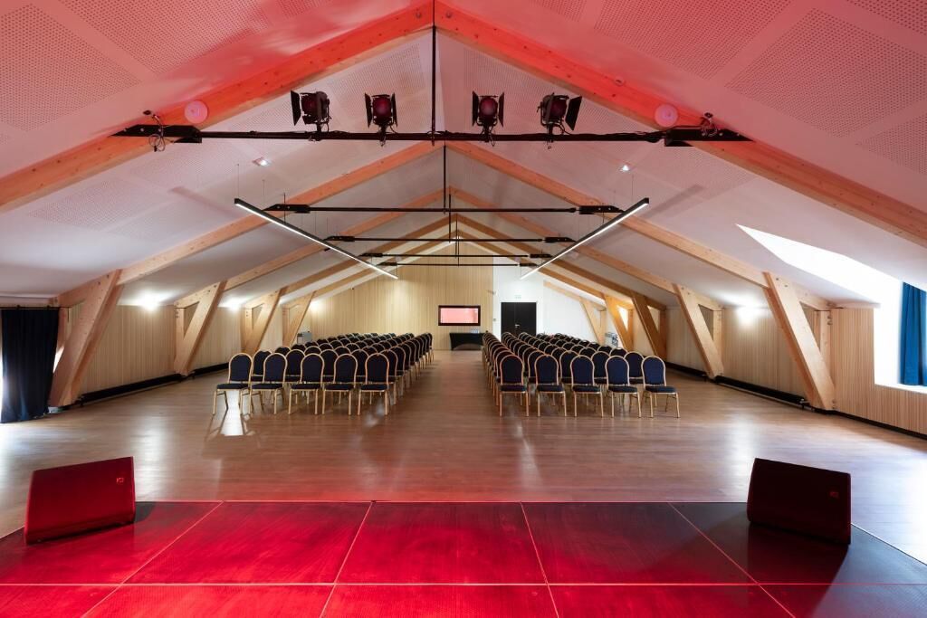 Un espace intérieur doté d’un plafond haut, de poutres en bois, de rangées de chaises et d’une scène.