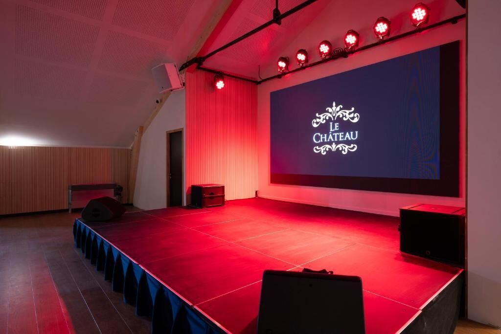 Een podium met een rode loper en een scherm waarop 'LE CHATEAU' staat, in een kamer met houten lambrisering en een schuine plafond.