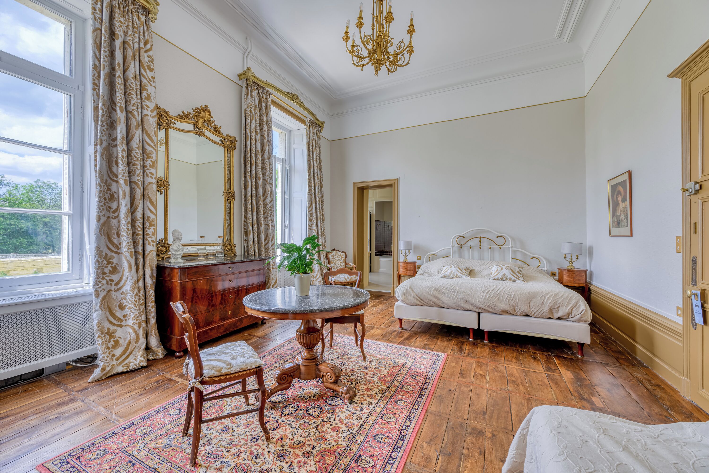 Une chambre à coucher avec un lit, une table ronde avec des chaises, un miroir et une fenêtre avec des rideaux.