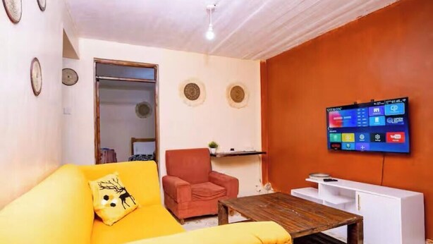 Un salón con sofá amarillo, un sillón rojo, una mesa de centro de madera, un mueble blanco y un televisor de pantalla plana montado en la pared.