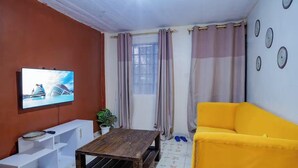 Una sala de estar con televisor, un sofá amarillo, una mesa de centro de madera y una ventana con cortinas.