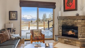 Una sala de estar acogedora con chimenea, un sofá, una silla y vistas a montañas a través de amplios ventanales.