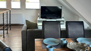 Moderni olohuone, jossa on ruokapöytä kahdelle, sohva, televisio ja portaat.