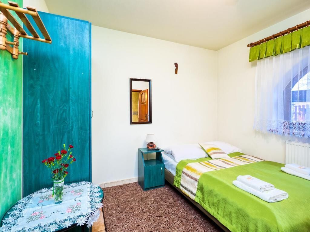 A bedroom with a bed, a nightstand, a mirror, a window with curtains, and a vase with flowers.