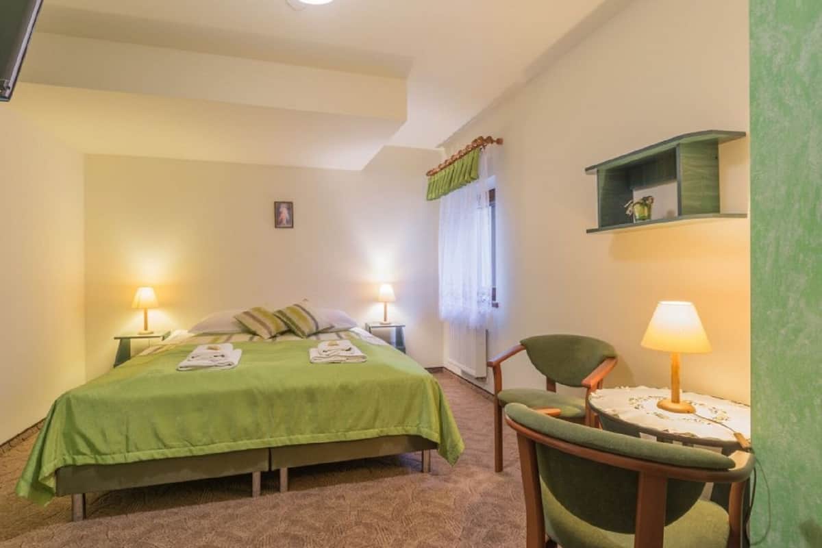 A bedroom with a bed, two bedside tables with lamps, a chair, a small table with a lamp, a window with curtains, and a green shelf.