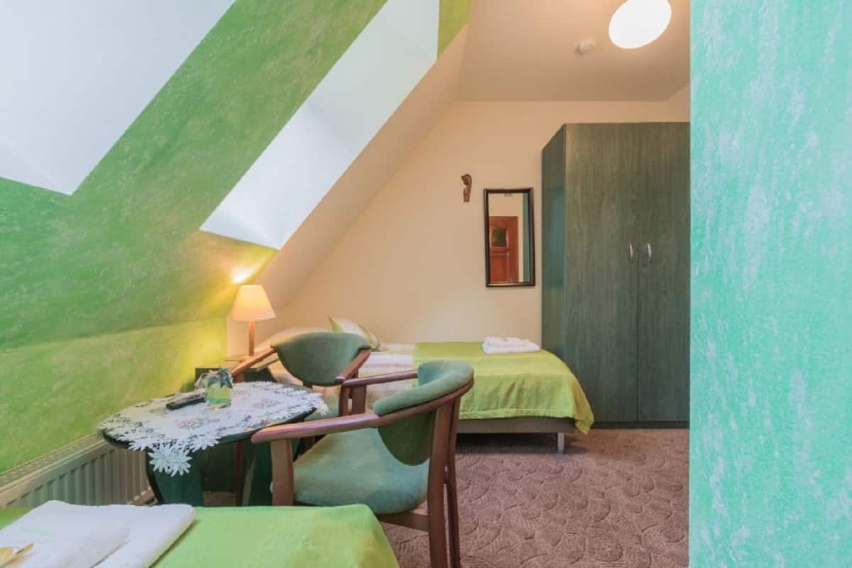 A small, cozy room with a sloped ceiling, a bed, a chair, a table with a lamp, and a wardrobe.