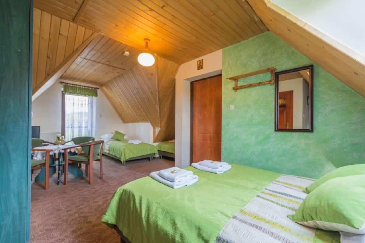 A room with two beds, a dining area with chairs, a wooden ceiling, and a green wall.