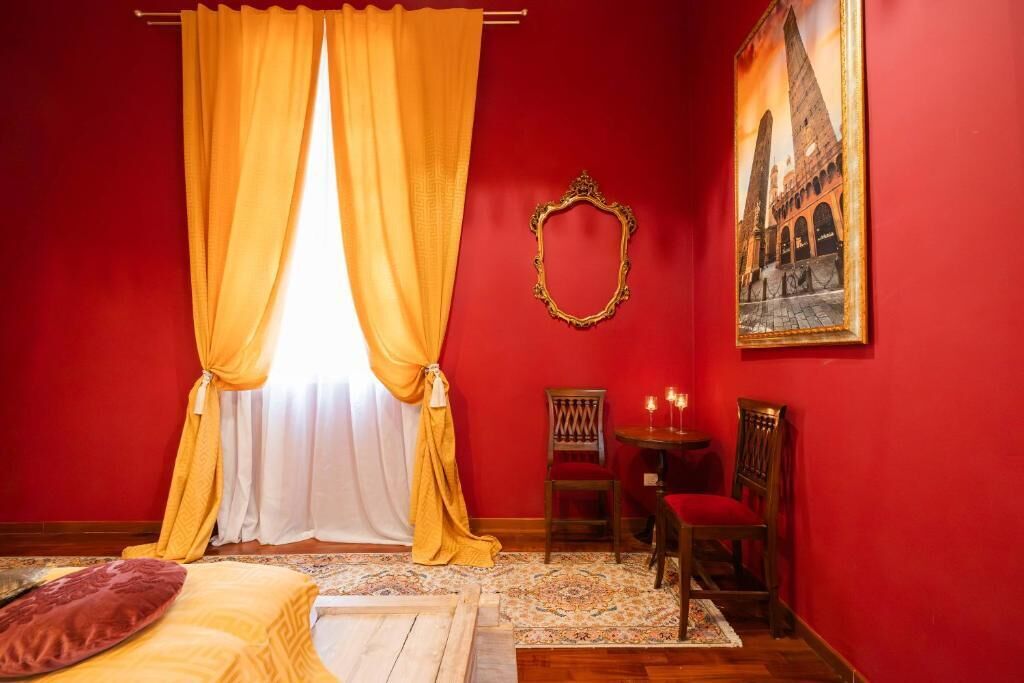 Een kamer met rode wanden, een bed met een rood kussen, een stoel, een kleine tafel met kaarsen, en een lijst met een afbeelding aan de muur.