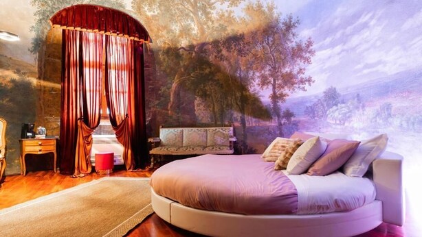 A bedroom with a round bed, a red curtain, a nightstand, a chair, and a wall mural. - Villa Meraville (Bologna)