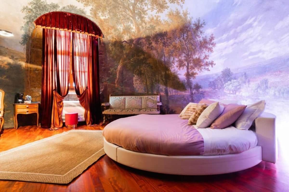 A bedroom with a round bed, a red curtain, a nightstand, a chair, and a wall mural.