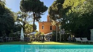 A clear swimming pool with lounge chairs and umbrellas, surrounded by trees and a building. - Villa Meraville (Bologna)