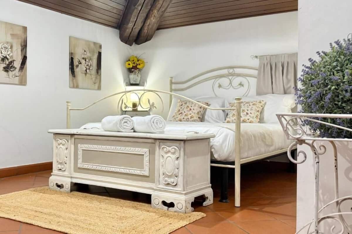 A bedroom with a bed, a bedside table with a lamp, a small rug, and a wall-mounted shelf with flowers.