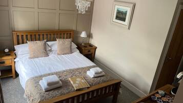 A hotel room with a bed, bedside tables, a nightstand, a wooden bench, a chandelier, a framed picture, and a tray with items.
