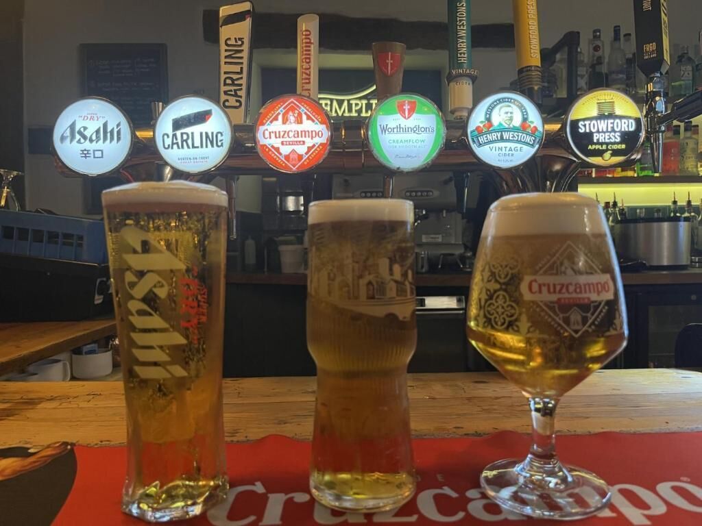 Three glasses of beer with labels from different brands, a bar counter, and a background with various bottles and a sign.