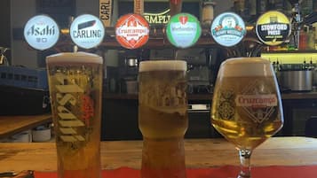 Three glasses of beer with labels from different brands, a bar counter, and a background with various bottles and a sign.