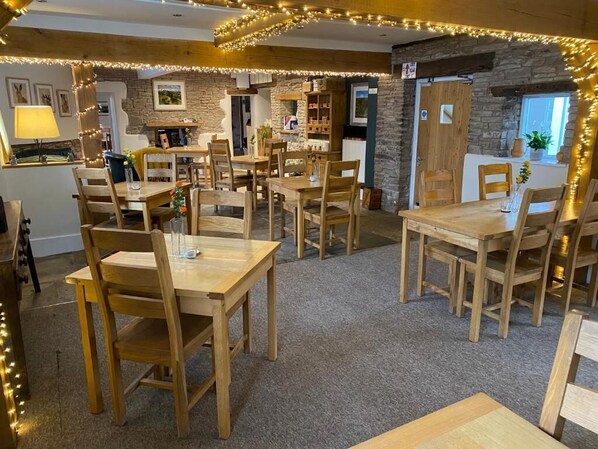A cozy dining area with wooden tables and chairs, stone walls, and string lights.