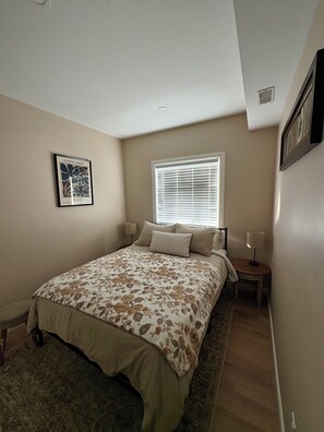 Une chambre à coucher avec un lit, une fenêtre avec des stores, une table de chevet avec une lampe et un tableau encadré au mur.