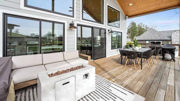 A modern outdoor living space with a wooden deck, a built-in fire pit, a dining area, and large sliding glass doors. - Lakefront - Private Dock - Near Ark - Sleeps 14 (Williamstown)