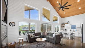 A spacious living room with a large gray sectional sofa, a round coffee table, and a view of a house through large windows. - Lakefront - Private Dock - Near Ark - Sleeps 14 (Williamstown)