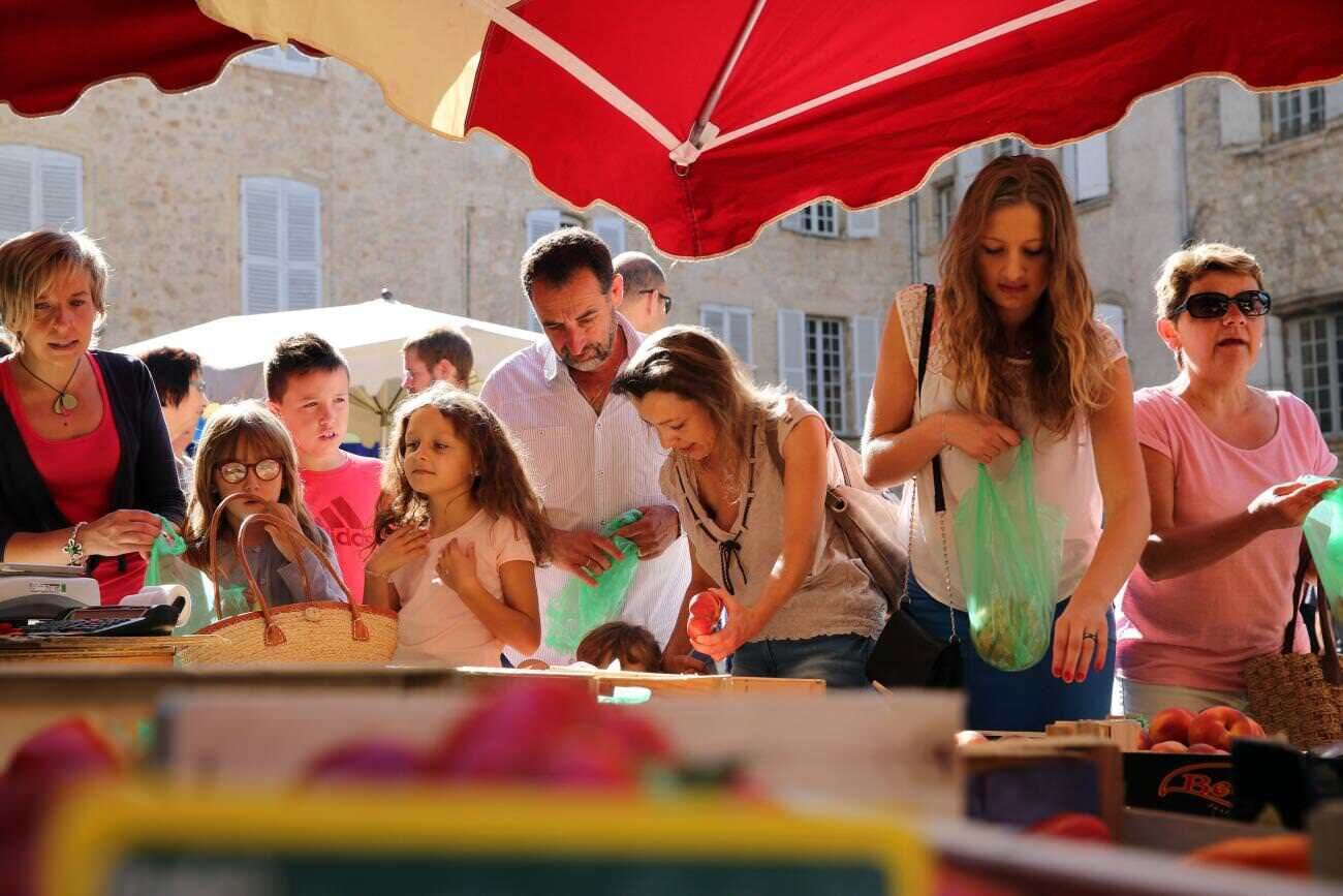 Un grup de persones a un mercat a l'aire lliure, amb un paraigua vermell, una taula amb productes i un edifici de pedra al fons.