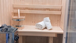 A sauna room with a bench, a metal container with rocks, and a glass container with a metal lid. - Apartment Kaiserperle at Haus Urvasi - Seaside-Flair with sauna and fireplace (Lübeck)
