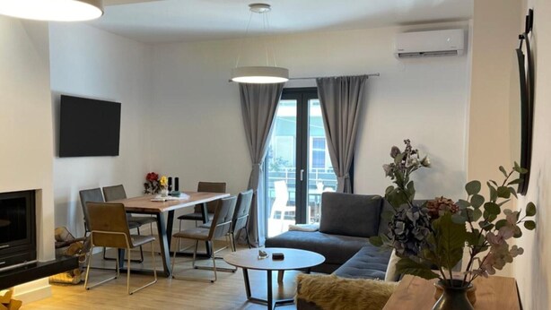 Ein modernes Wohnzimmer mit einer Couch, einem Couchtisch, einem Essbereich mit Stühlen, einem an der Wand montierten Fernseher und einem großen Fenster mit Vorhängen.