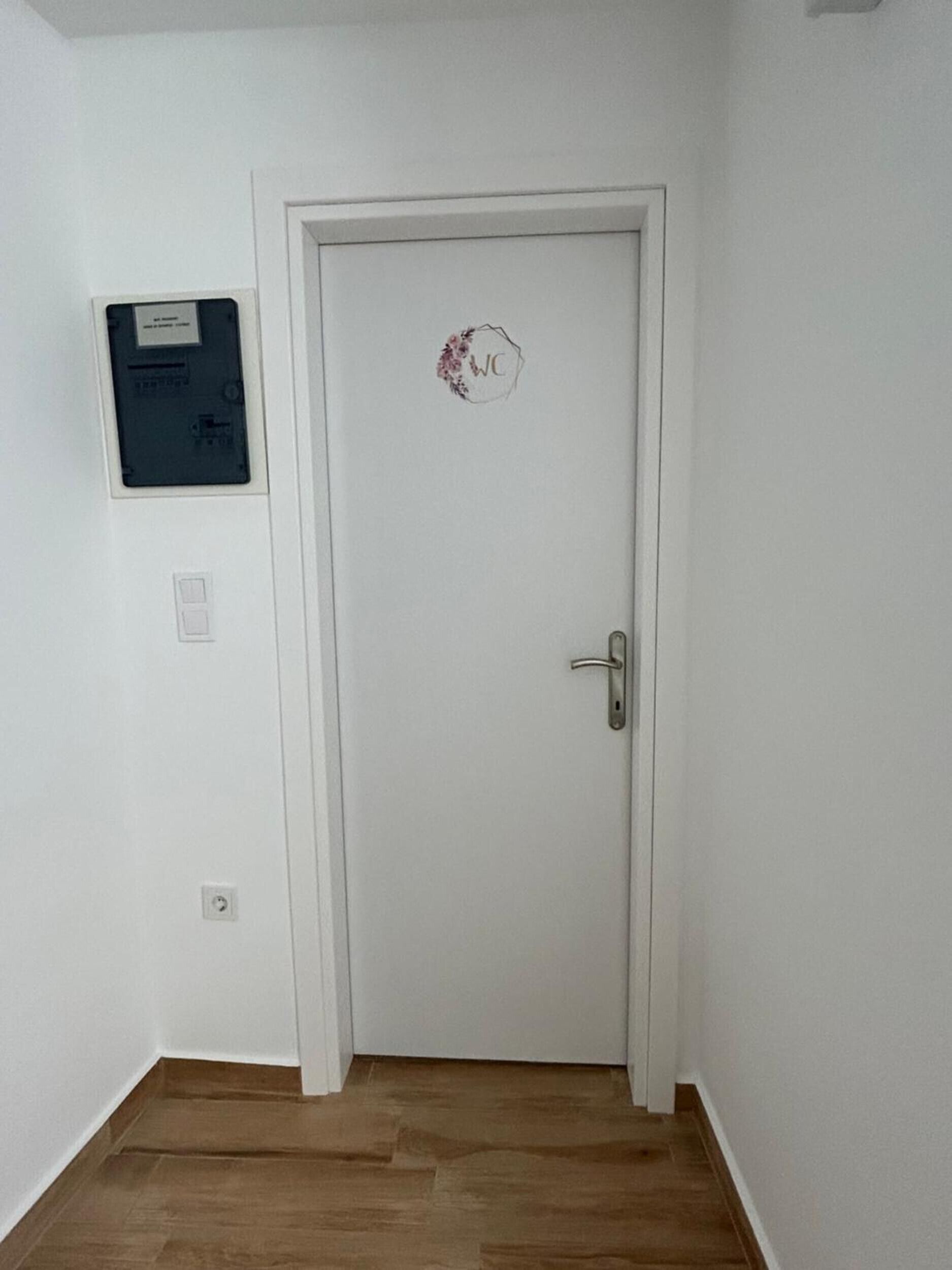Una puerta blanca con cerradura y un adhesivo circular, un cuadro eléctrico empotrado en la pared, y un interruptor de luz.