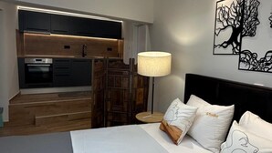 Habitació d'hotel moderna amb un llit, una tauleta de nit, una làmpada, una obra d'art a la paret i una cuina visible a través d'una porta oberta.
