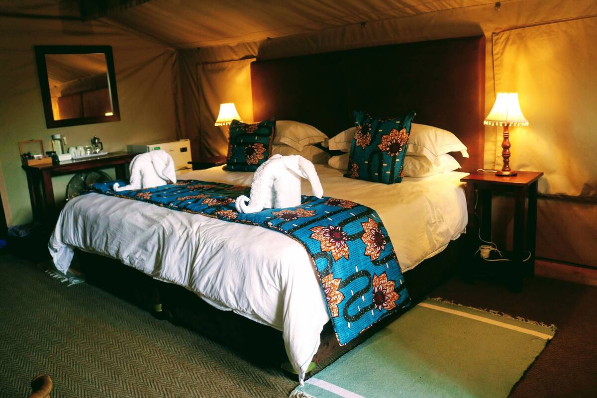 A double bed with a blue floral bedspread, two pillows, a bedside table with a lamp, and a mirror on the wall.