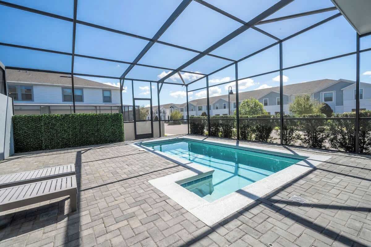 A swimming pool with a glass enclosure, surrounded by a paved patio and a hedge.