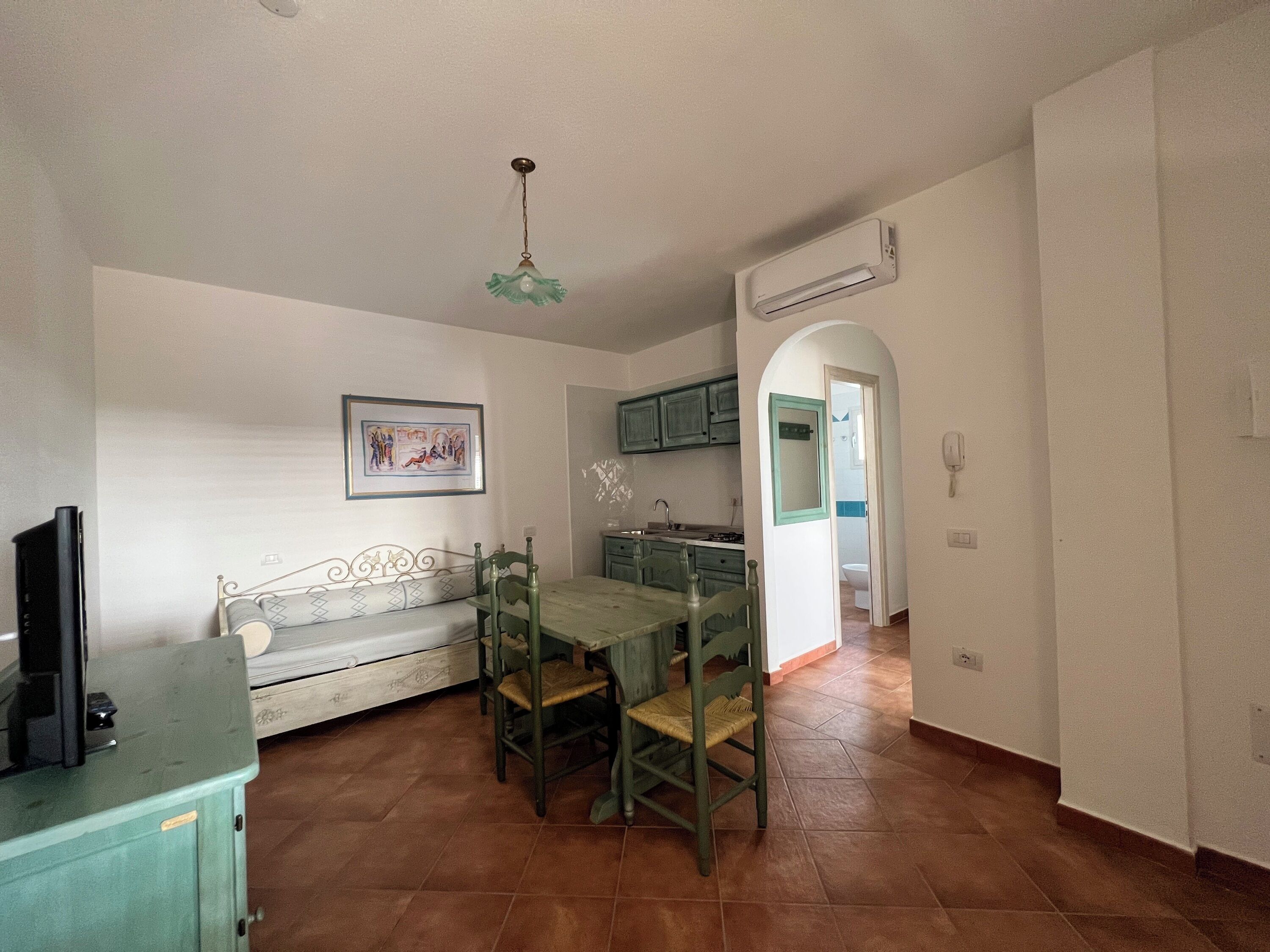A dining area with a wooden table and chairs, a bench, a ceiling light fixture, and a kitchen area with cabinets and an air conditioner.